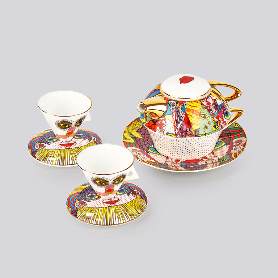 A porcelain teapot and two matching cups with saucers, decorated with a colorful clown face illustration and gold trim, set against a solid gray background.