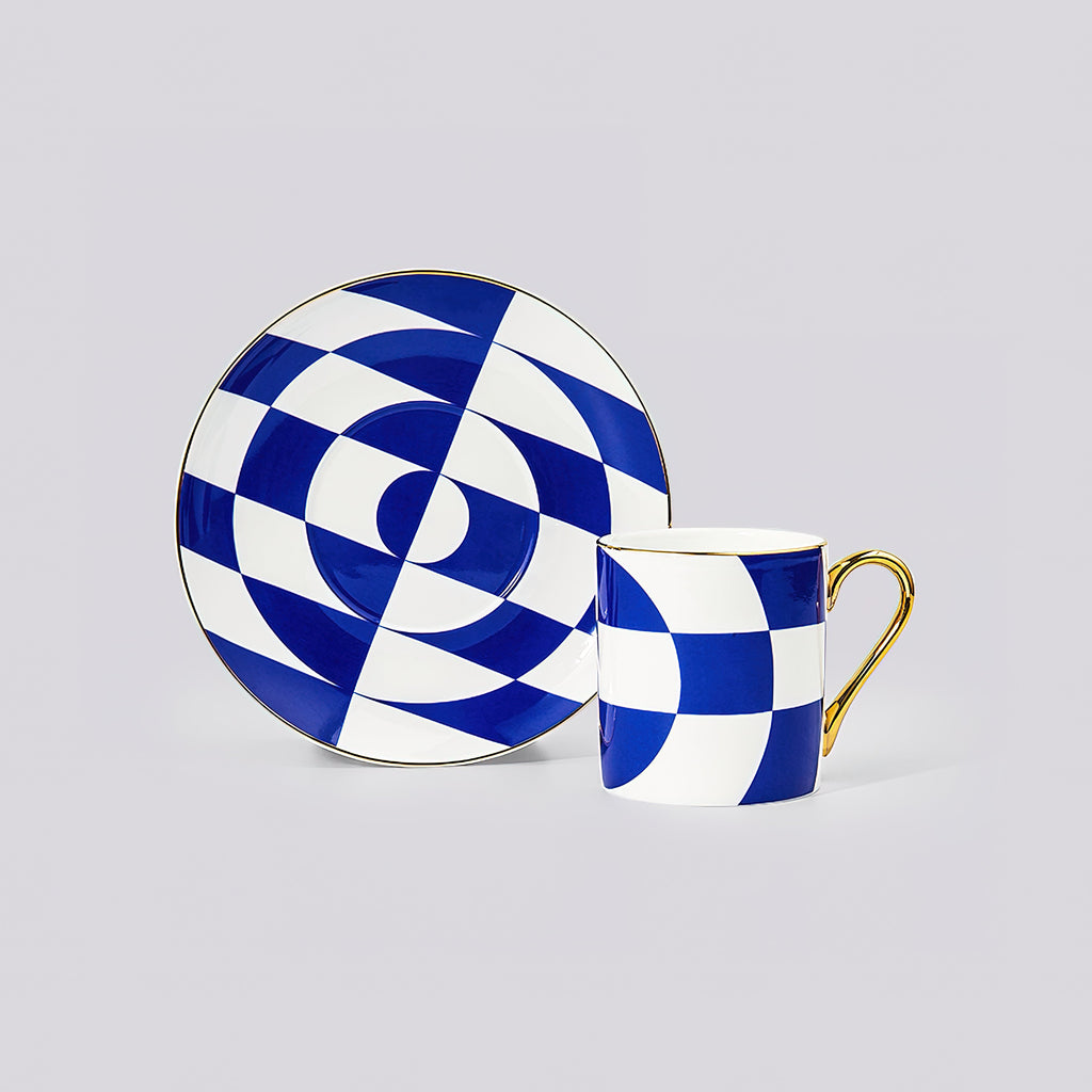 A blue and white geometric checkered cup and saucer set with gold trim, displayed on a neutral light gray background.
