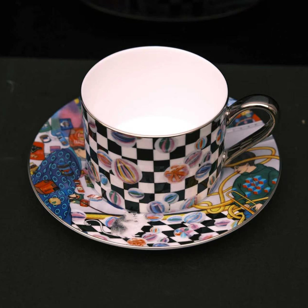 A porcelain teacup and saucer set featuring a black and white checkerboard pattern on the cup exterior and a vibrant, multicolored macaroni illustration on the saucer, set against a dark background.