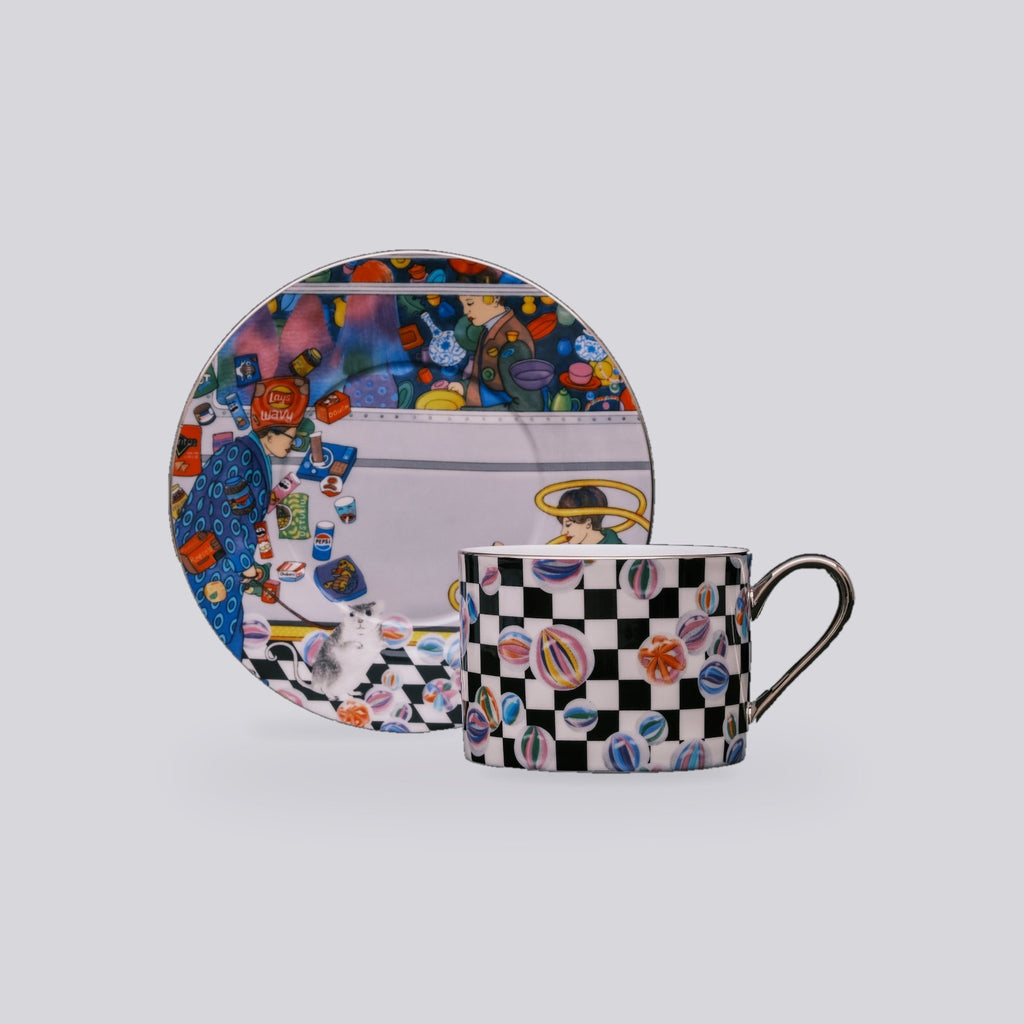 A matching ceramic cup and saucer set with a colorful cosmic-themed print and a black-and-white checkered pattern with multicolored planet motifs, shown on a light gray background.