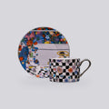 A matching ceramic cup and saucer set with a colorful cosmic-themed print and a black-and-white checkered pattern with multicolored planet motifs, shown on a light gray background.