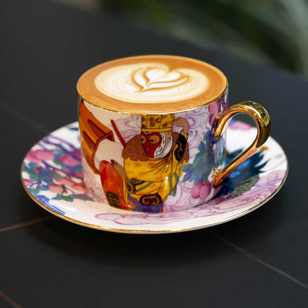 A decorated ceramic cappuccino cup and saucer set on a dark table, featuring a colorful Art Nouveau style illustration with gold trim and a heart latte art design on top.
