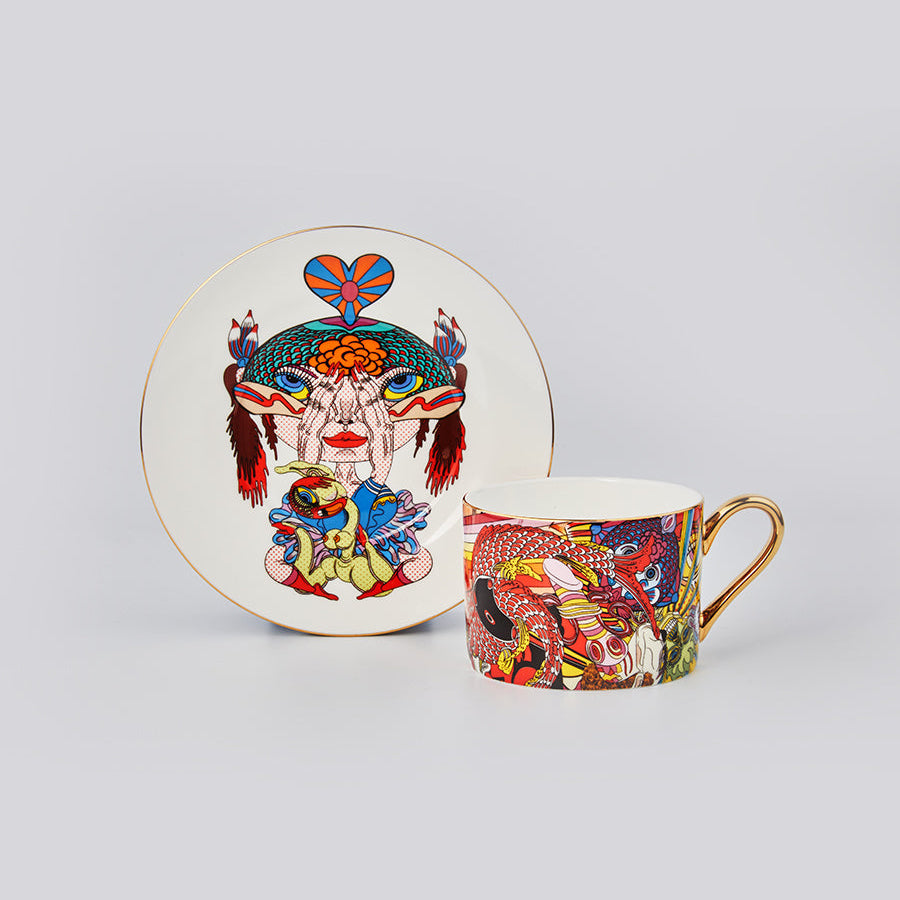 A matching ceramic cup and saucer set featuring a vibrant, detailed multicolor illustration of a stylized face with a heart-shaped headpiece, on a light gray background.
