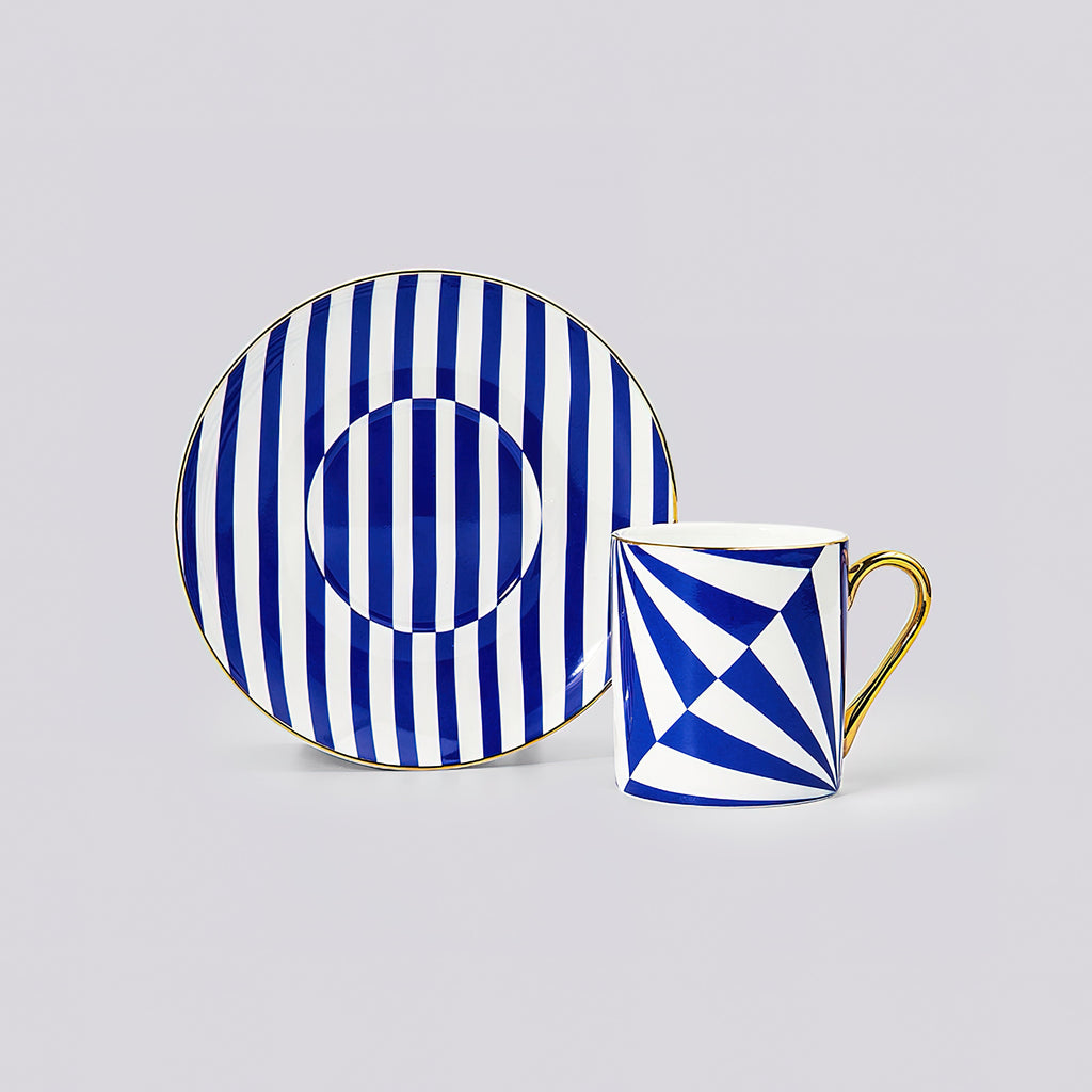 A blue and white patterned ceramic cup and saucer set on a light gray background. The saucer features vertical blue and white stripes with a circular motif, while the cup has a geometric blue and white pattern with a gold handle.