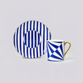A blue and white patterned ceramic cup and saucer set on a light gray background. The saucer features vertical blue and white stripes with a circular motif, while the cup has a geometric blue and white pattern with a gold handle.