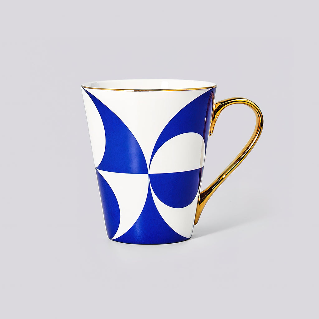 A white ceramic mug featuring a bold blue geometric pattern and a polished gold handle, set against a solid light gray background.