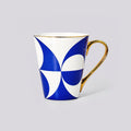 A white ceramic mug featuring a bold blue geometric pattern and a polished gold handle, set against a solid light gray background.