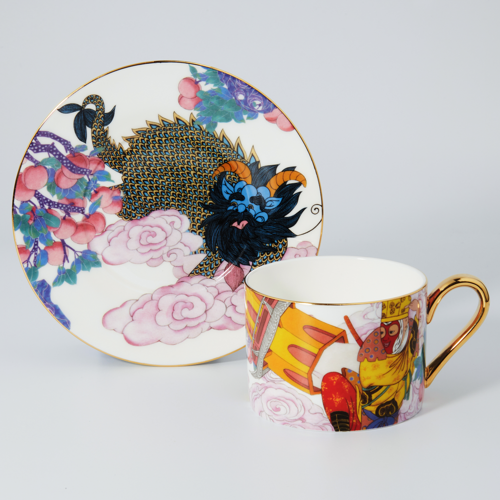 A porcelain cup and saucer set with intricate hand-painted illustrations, including a black and gold dragon, pink clouds, and colorful floral motifs, accented with gold trim on a light gray background.