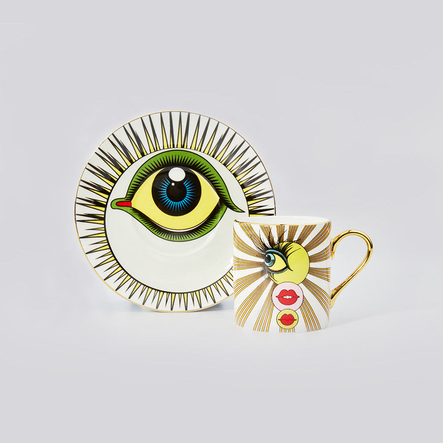 A ceramic mug and matching saucer featuring a stylized eye illustration with radiating gold stripes on a white background, photographed on a light gray studio backdrop.