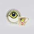 A ceramic mug and matching saucer featuring a stylized eye illustration with radiating gold stripes on a white background, photographed on a light gray studio backdrop.