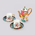A porcelain tea or coffee set on a light gray background, featuring a teapot with a colorful illustrated body, gold-tone handle and spout, and two matching cups and saucers with gold rims and abstract multicolor patterns.